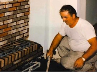 A man with a tool in his hand kneels near a brick wall.