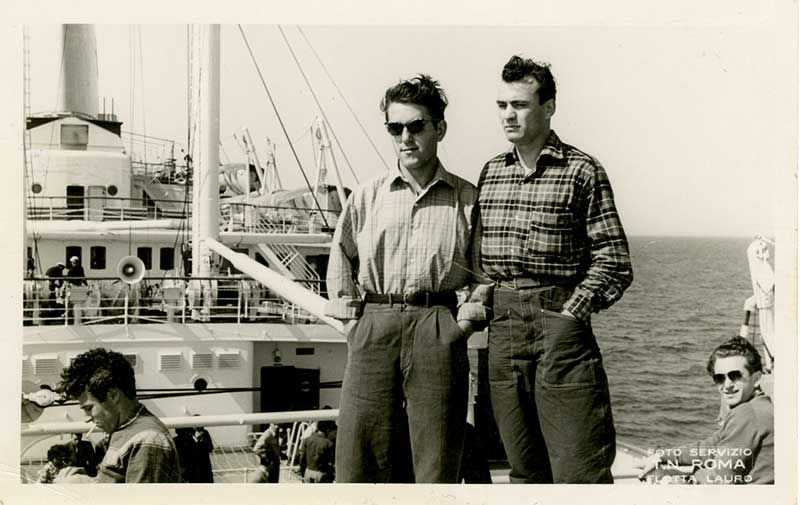 Two young men stand next to each other on a ship’s deck.