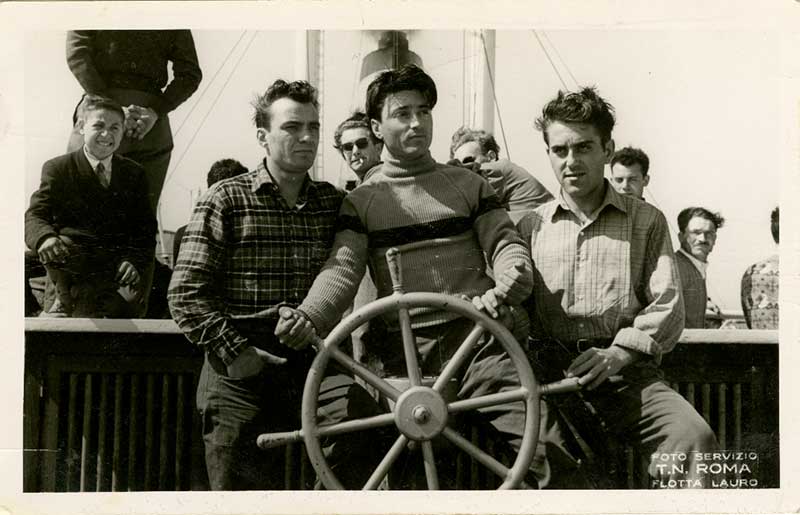Three young men stand behind a ship’s steering wheel and pretend to steer.