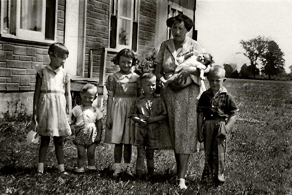 A young woman stands on the grass with five small children and a baby.