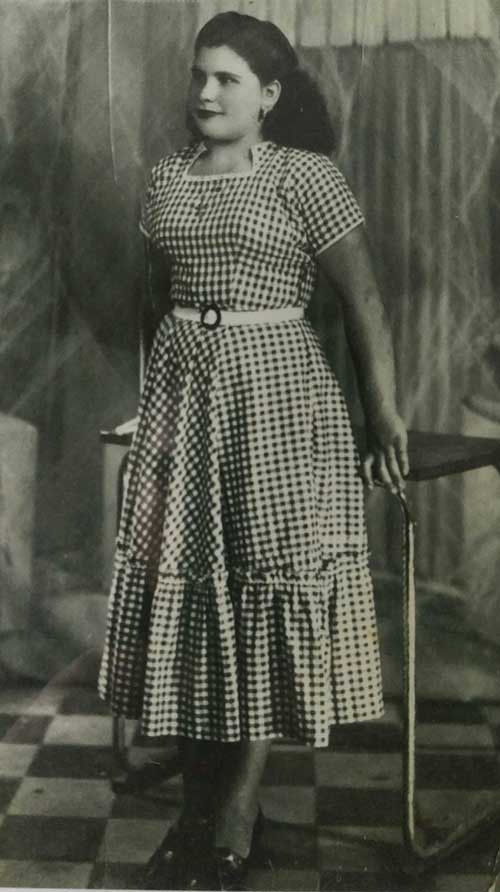 A young woman with her hair pinned back stands against a table and wears a checkered dress.