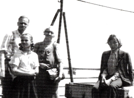 Four people on deck on a ship, pulleys in the background.