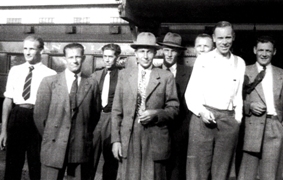 Old photograph of several men standing in suits, ties and hats.