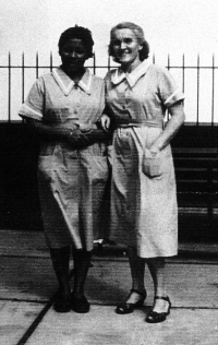 Two women dressed as nurses, standing in front of an iron fence.