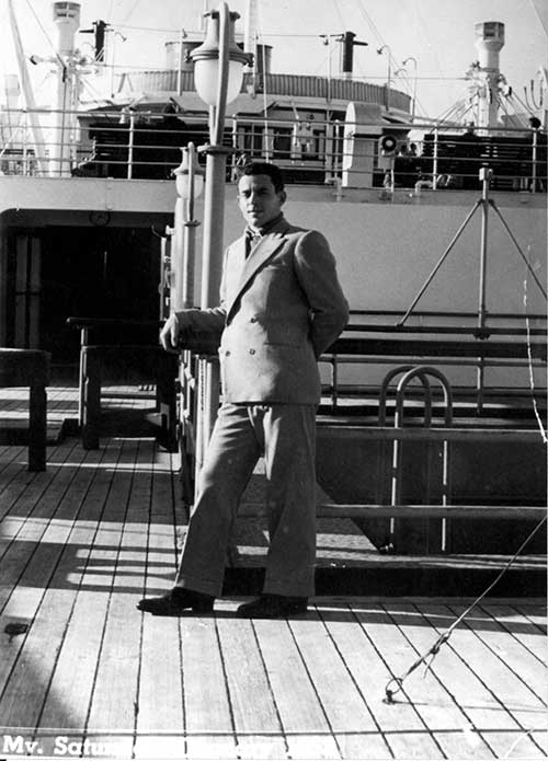 A young man wearing a suit is leaning on a ship’s railing.
