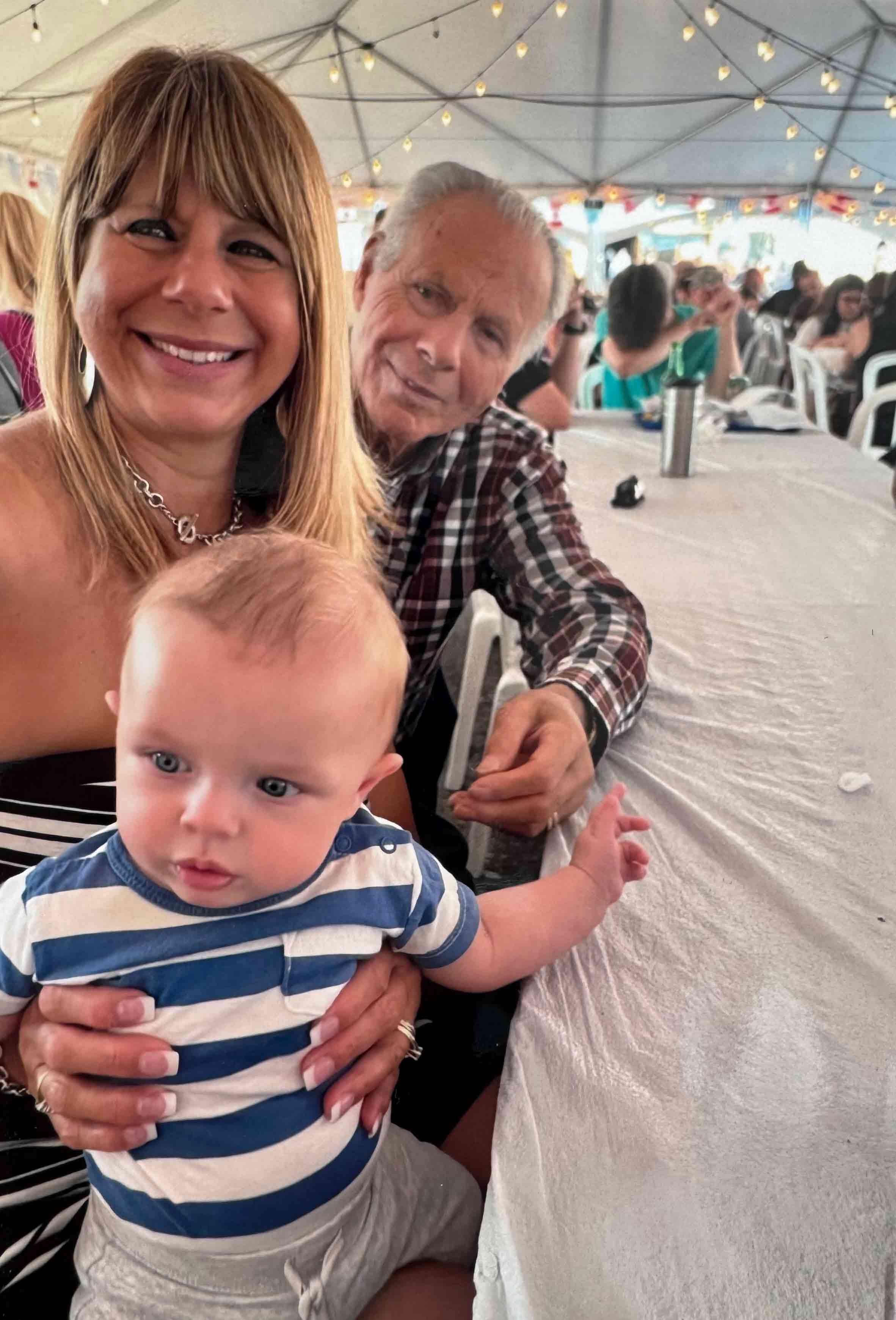 A smiling Greek man, woman, and baby sit at a long table.