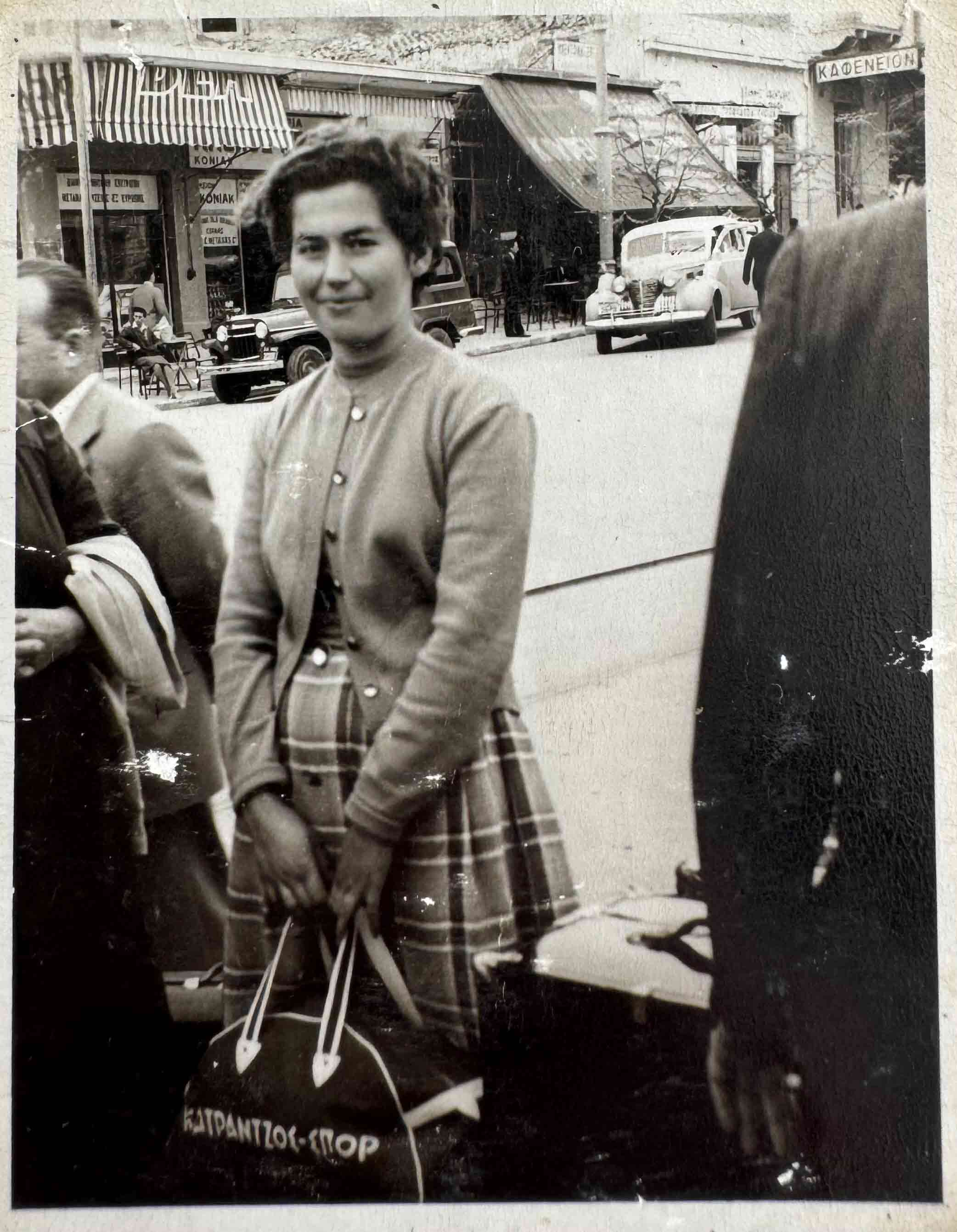 A black and white photo of a Greek lady holding luggage and smiling.