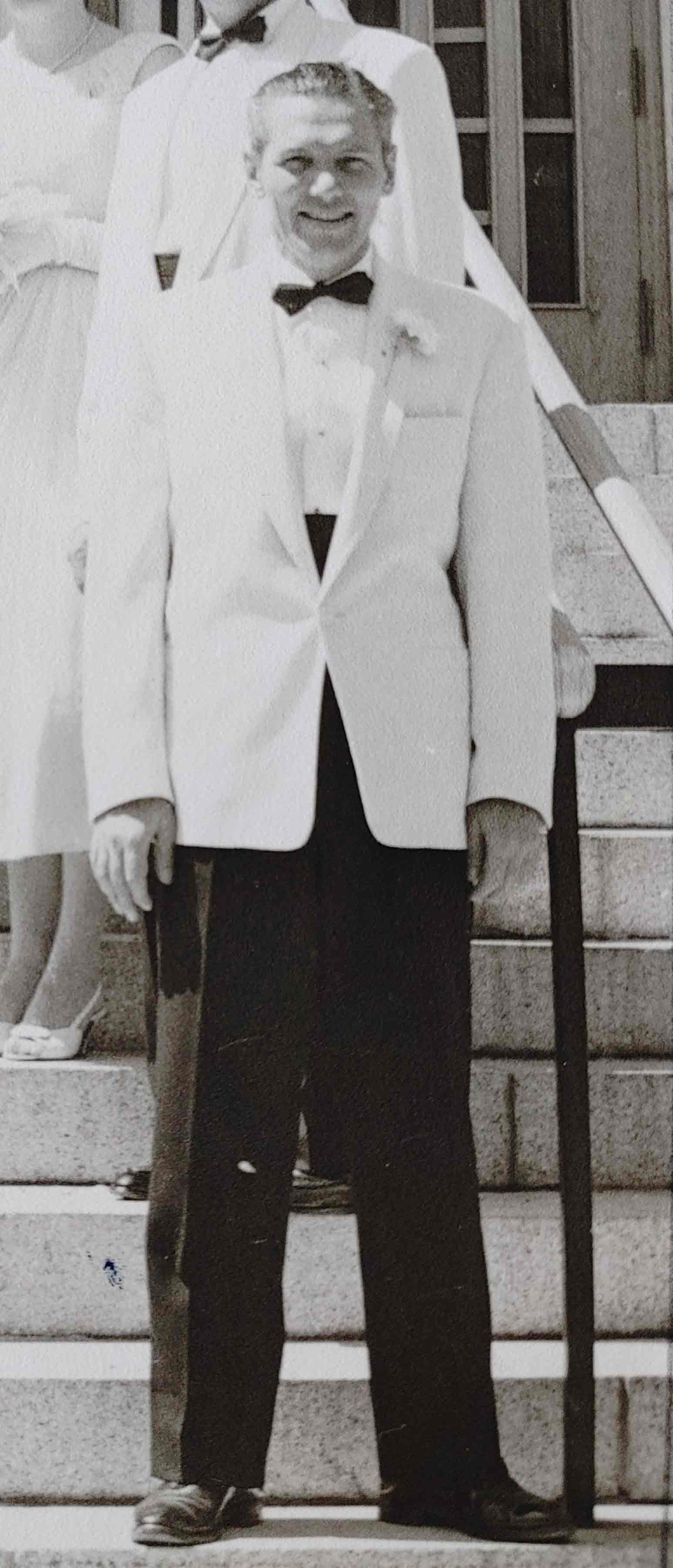 A black and white photo of a white man in black pants and a white suit jacket, standing on stairs with others behind him.