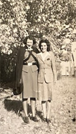 Sepia photo of two white women dressed in uniform, standing outside with arms around each other.