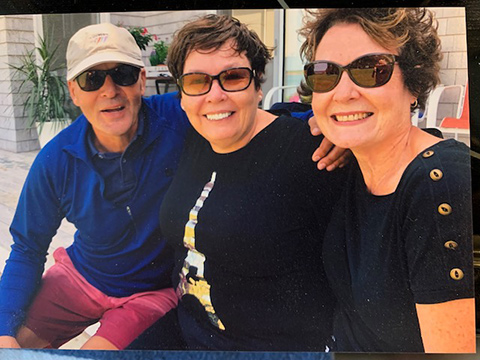 Two white women and one man in sunglasses, sitting on a deck and smiling at the camera.