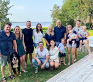 A group of ten white adults, three children, and one large dog on the grass in front of the water.