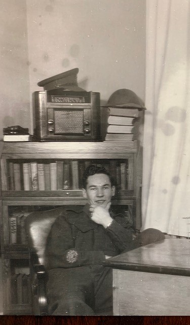 Black and white photo of a smiling white man, chin in his hand, seated at a desk with a bookcase behind him.