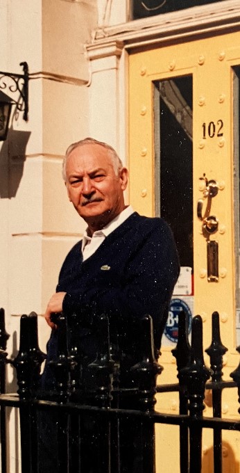 An older white man outside a front door, with one arm leaning on a black fence.