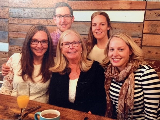 Four smiling white women and one man at a restaurant with wooden walls and drinks on the table.